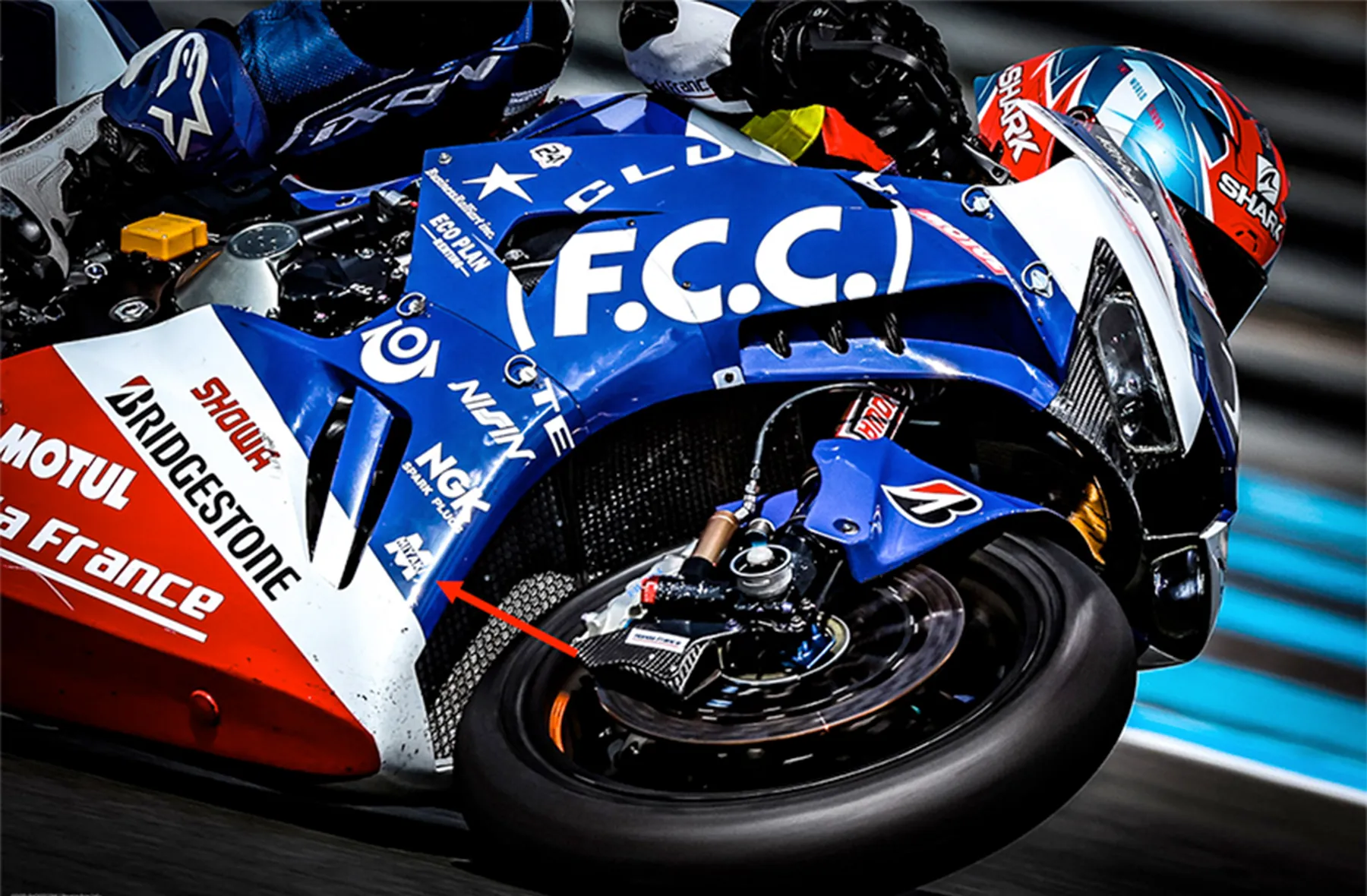 F.C.C. TSR racing motorcycle leaning into a track corner with an arrow highlighting the Miyaki logo.