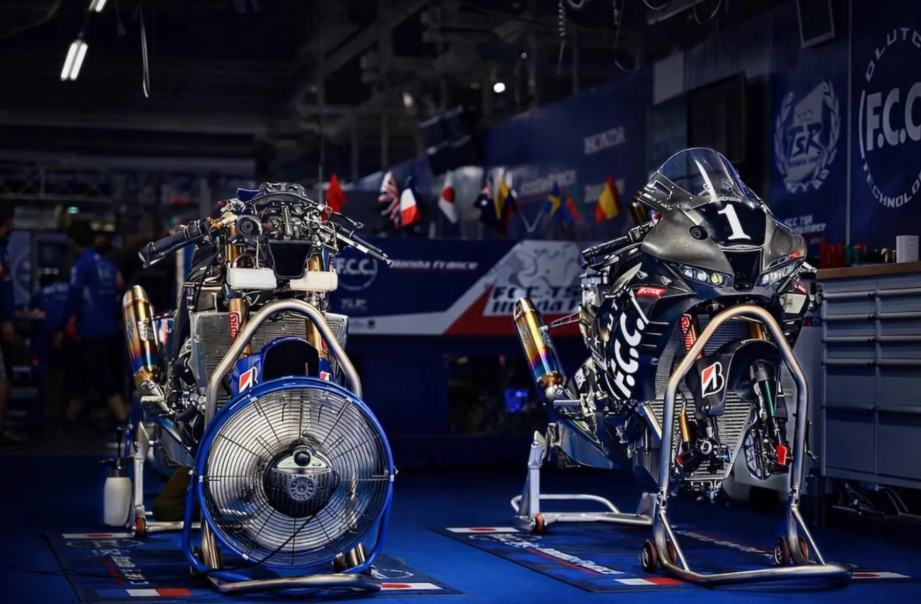 Two stripped-down TSR racing motorcycles resting on stands with large cooling fans in the pit garage.